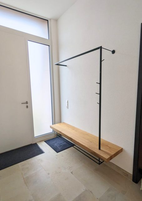 Minimalistische Garderobe mit Holzbank und schwarzem Metallgestell als Kleiderstange, in hellem Flur mit Glastür.