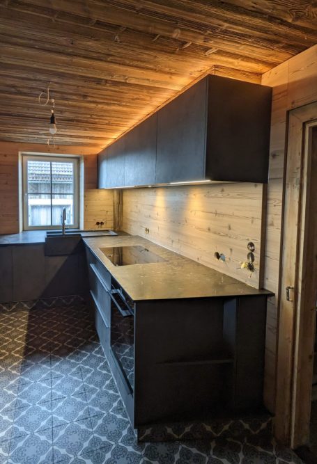 Detailansicht der modernen Küche mit schwarzen Schränken, heller Holzverkleidung an Wand und Decke sowie gemustertem Bodenbelag.