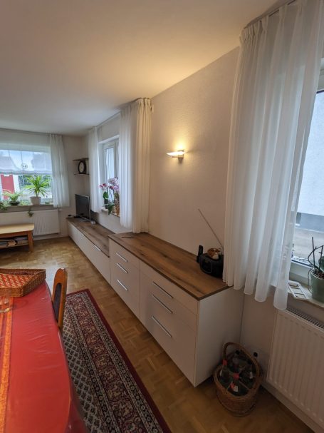 Langes Sideboard mit weißen Schubladen und Holzoberfläche in einem Wohnzimmer mit großen Fenstern und Vorhängen.