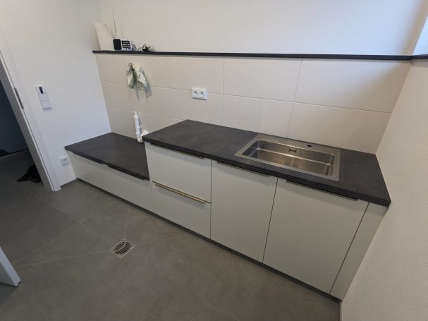 Hauswirtschaftsraum mit Einbauschränken in Hellgrau, schwarzer Arbeitsplatte und Edelstahlspüle vor weiß gefliester Wand.