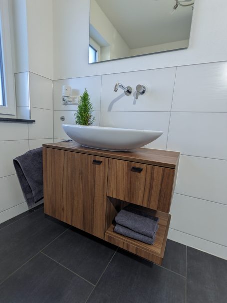 Badezimmer mit wandhängendem Holzunterschrank, großem ovalen Aufsatzwaschbecken und weiß gefliester Wand.