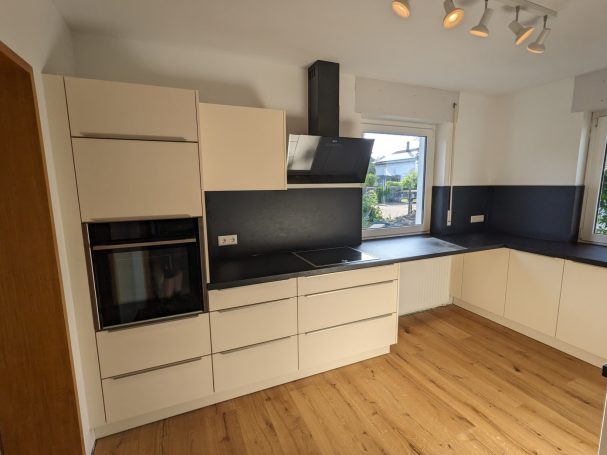 Moderne Küche mit weißen Schränken, schwarzer Arbeitsfläche, Dunstabzugshaube und großem Fenster, kombiniert mit hellem Holzfußboden.