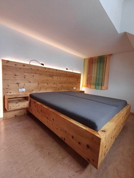 Schlafzimmer mit Bett aus massivem Zirbenholz, großem Kopfteil mit Beleuchtung und integrierten Nachttischen.