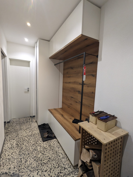 Flurgarderobe mit Holzrückwand, Sitzbank, schwarzer Kleiderstange und weißen Oberschränken.
