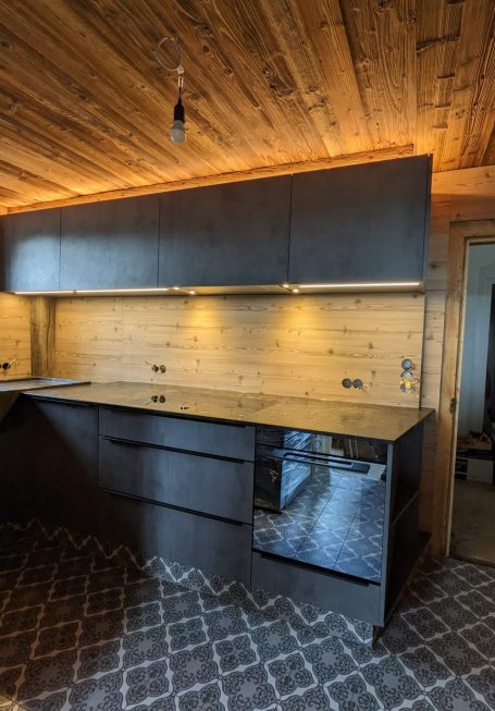 Moderne Einbauküche mit dunklen Fronten und Arbeitsplatte, Holzdecke und Holzrückwand, stilvolle Beleuchtung und großem Fenster.