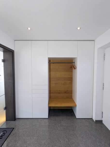 Weiße Einbau-Garderobe mit Holzrückwand, Sitzbank und offenen Kleiderhaken, kombiniert mit geschlossenem Stauraum.