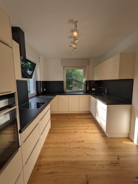 Helle L-förmige Einbauküche mit matten weißen Fronten, schwarzer Arbeitsplatte und Rückwand, Holzfußboden und großem Fenster.