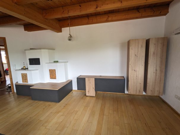 Wohnzimmermöbel aus Holz und anthrazitfarbenen Elementen mit Regalen und Bank auf Holzboden unter sichtbaren Deckenbalken.