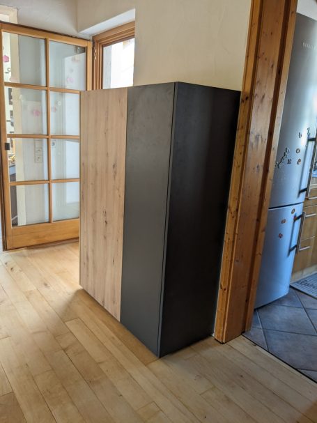 Moderner Schrank in Holz-Schwarz-Kombination, stehend vor einer Glastür im Innenbereich.